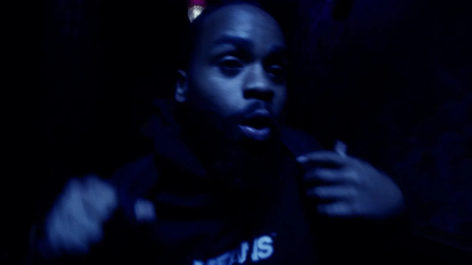 Dark blue-lit frame of a man in a hoodie mid-gesture, face blurred and lit from the side