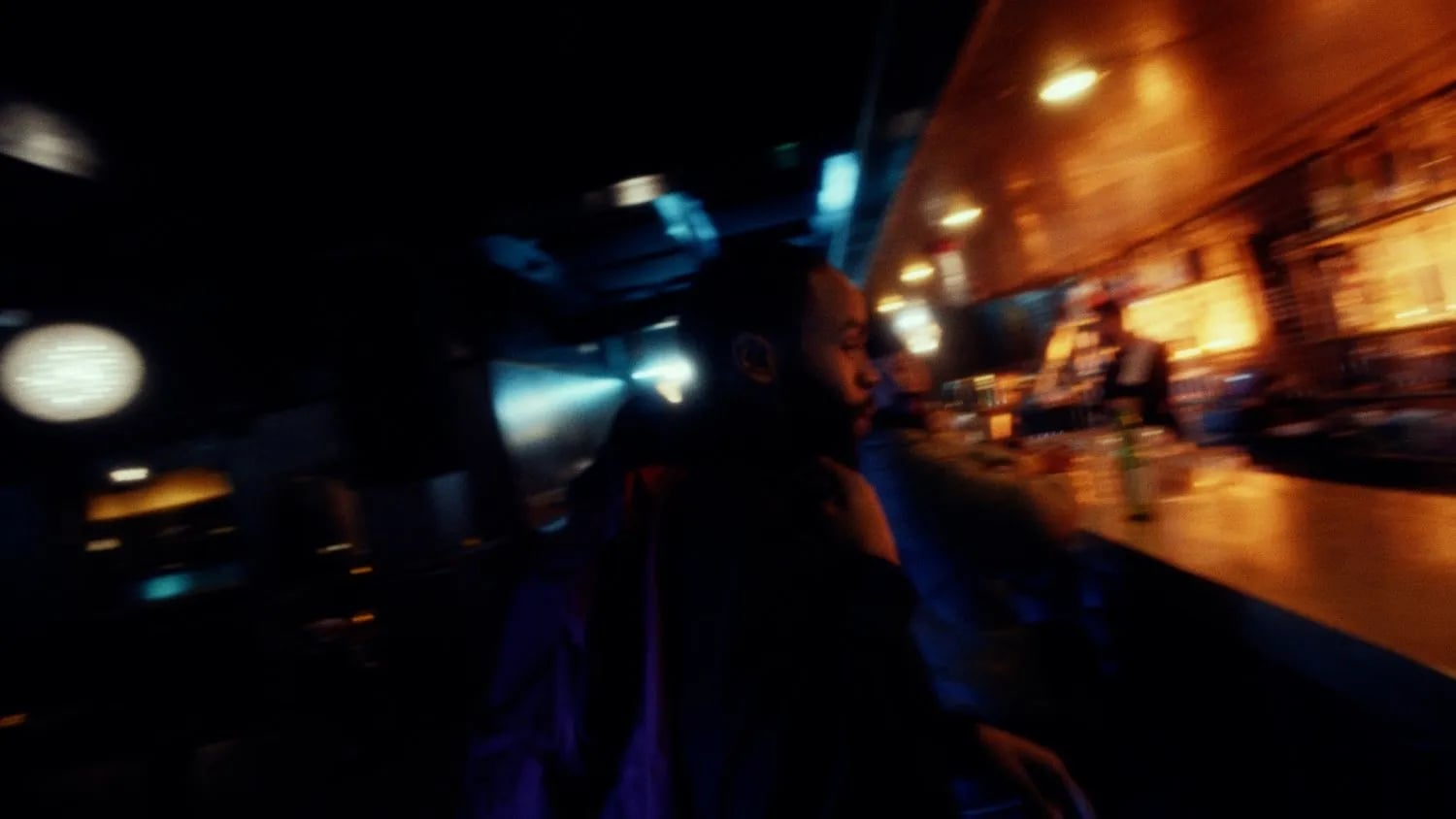 Frame from the No Dough music video: man at the end of a bar, lit orange, with blurred lights in a dark room