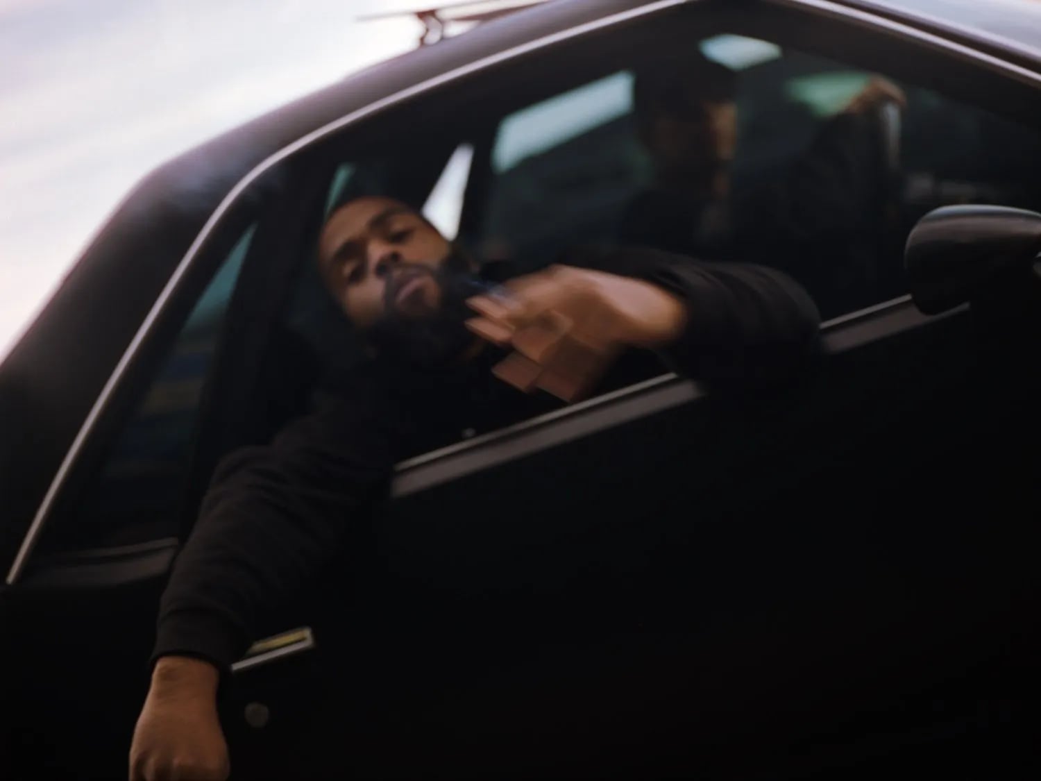 Blurred man with a beard leaning out of a dark car window with one hand raised, a second person behind him