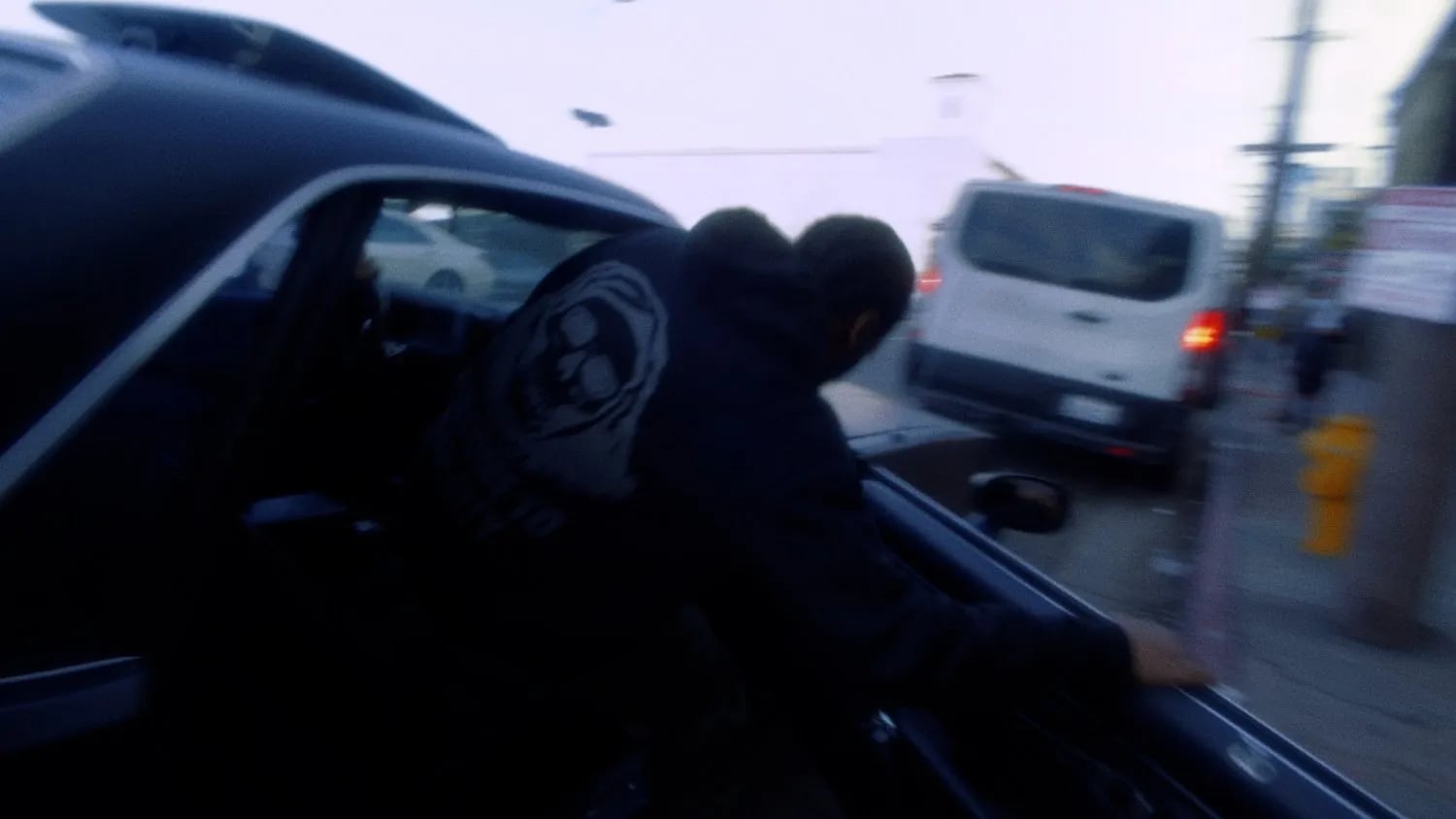 Frame from the No Dough music video: person in a black hoodie with a skull graphic leaning over a car door, white van behind