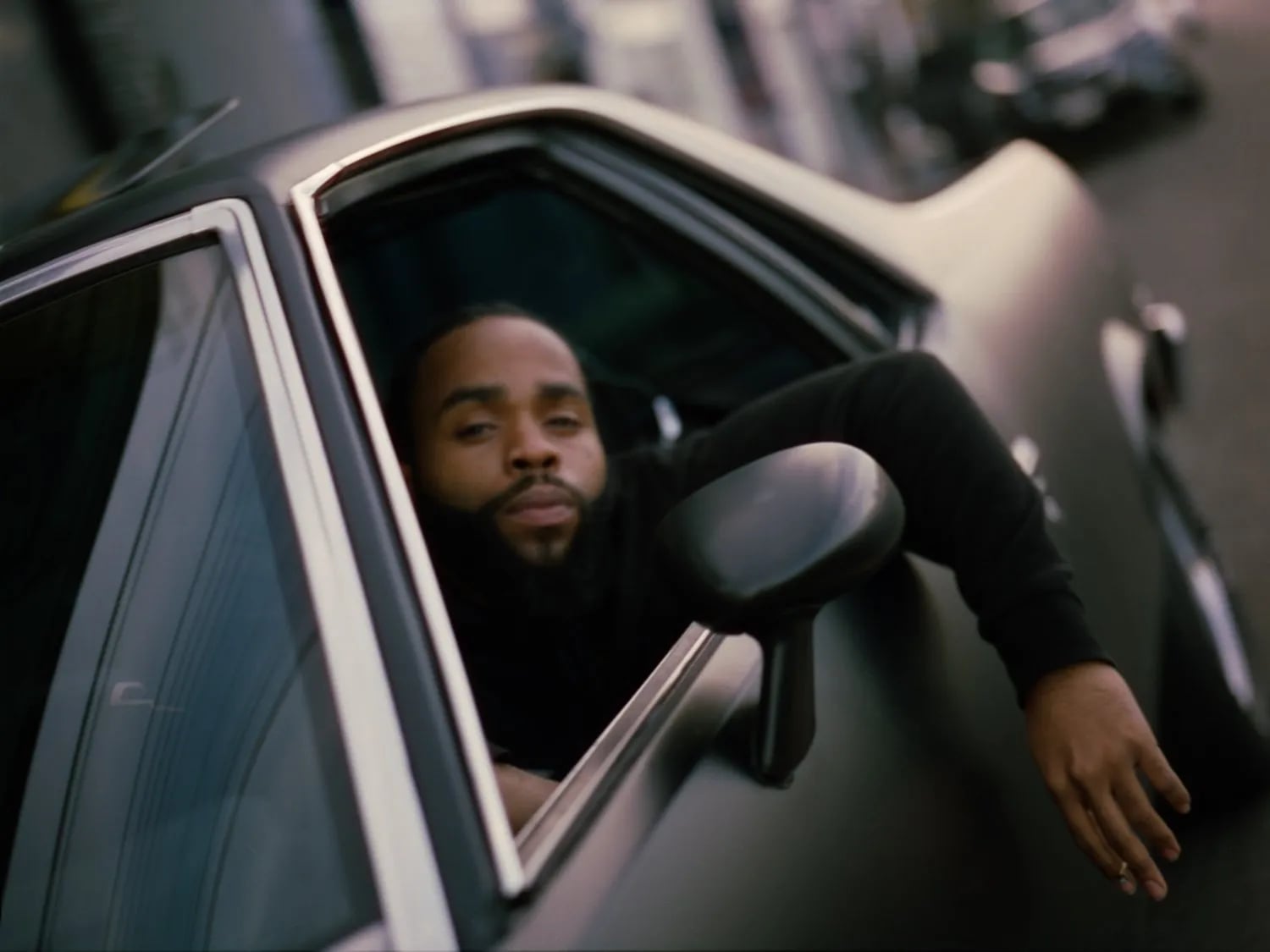 Bearded man in a black sweater reclining with his head and arm out of a matte black car window