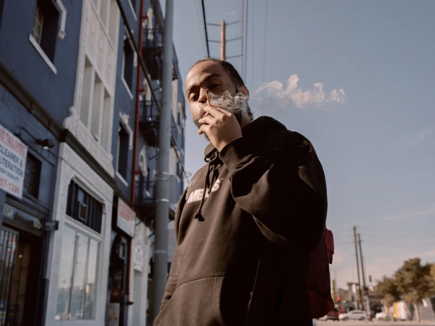 Man in a brown hoodie on a street exhaling smoke, blue buildings and power lines behind him