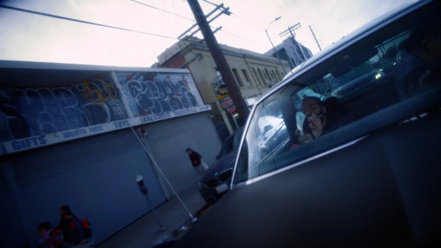 Man in a car seen through its window, graffiti-covered shop shutters and a parking meter on the street outside