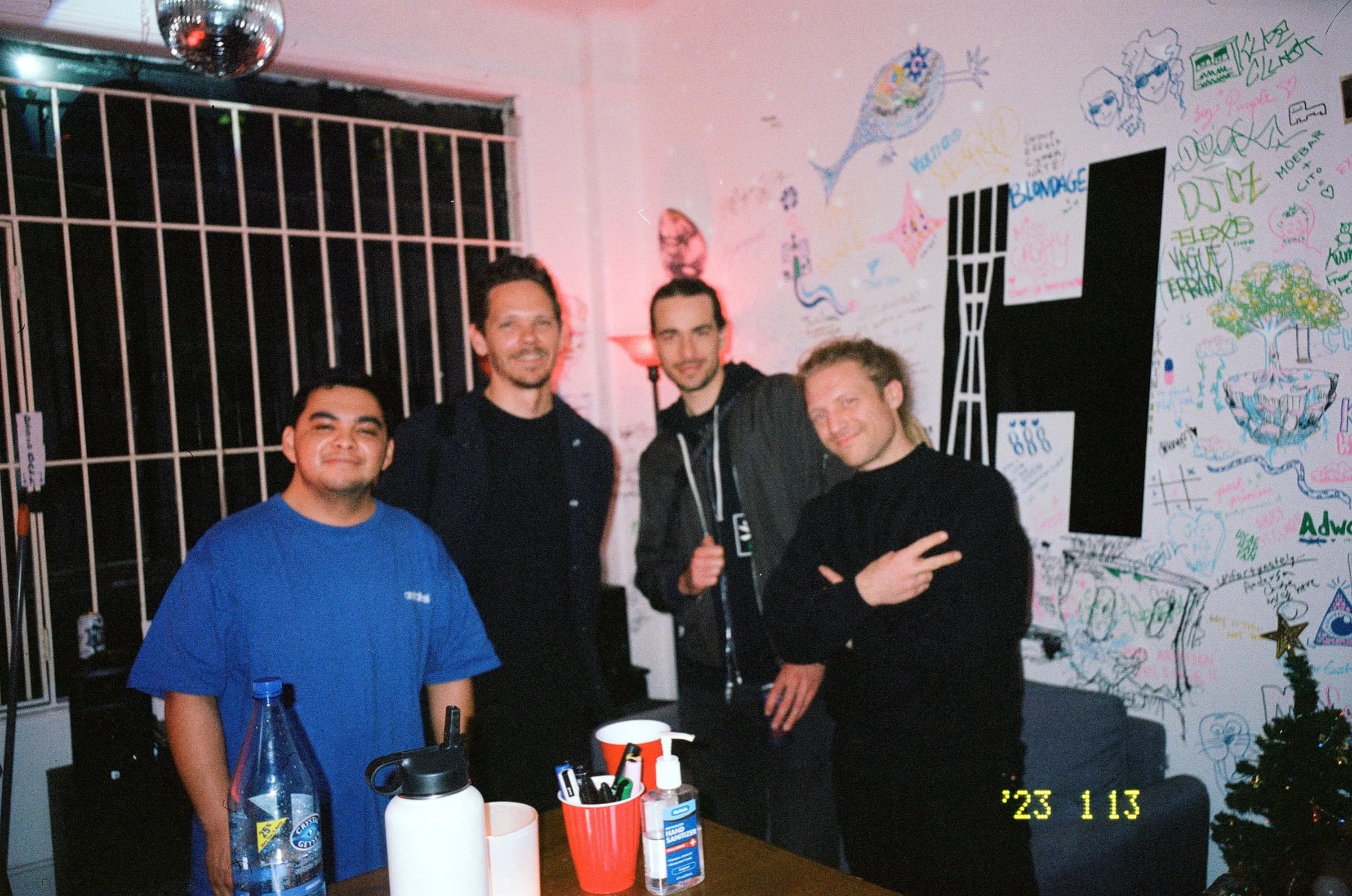 Skyler and three people posing at a table in a radio station whose wall is covered in marker drawings, with a disco ball above