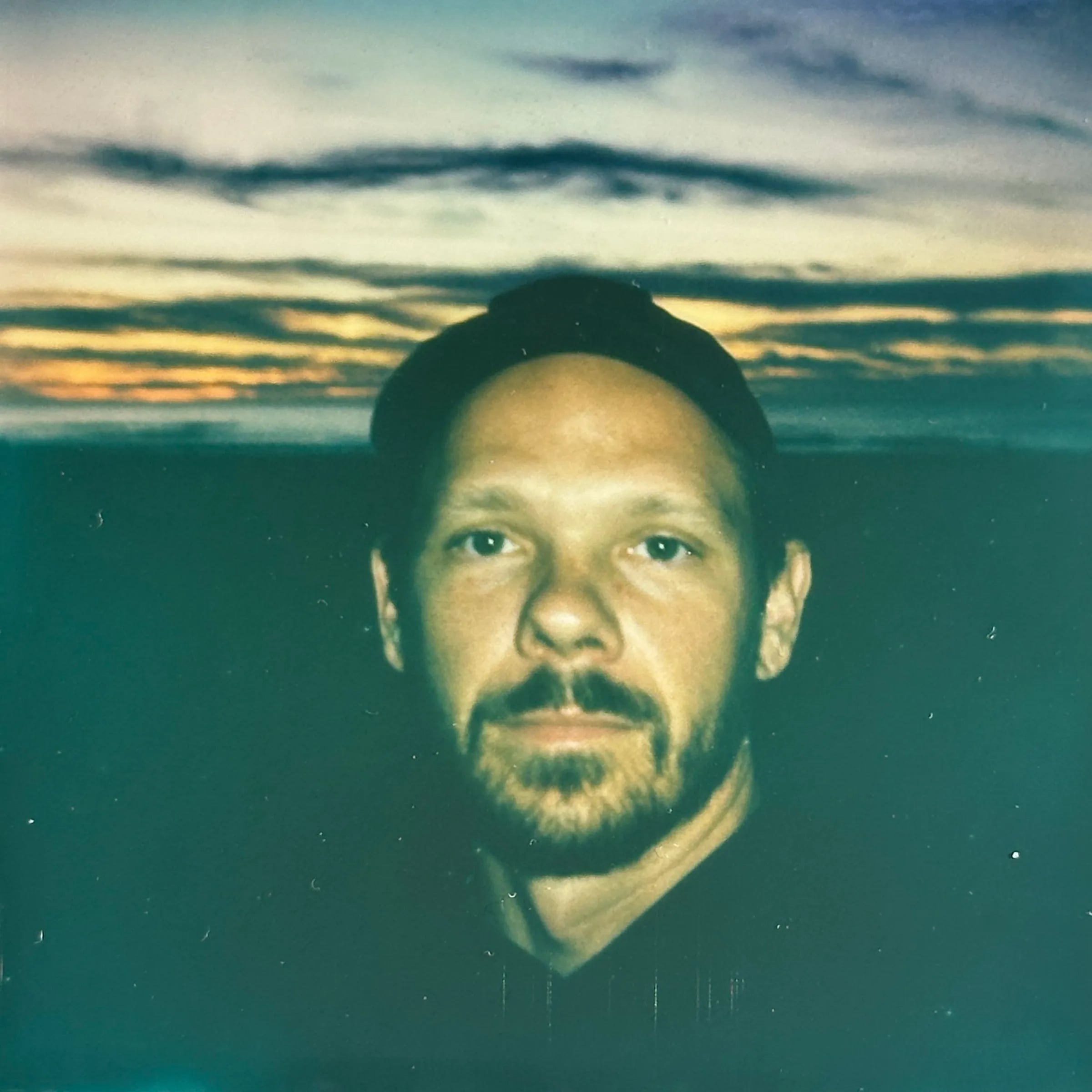 Close portrait of Skyler, bearded, in a dark cap on a beach against a dusky sunset with orange-lit clouds and teal tones