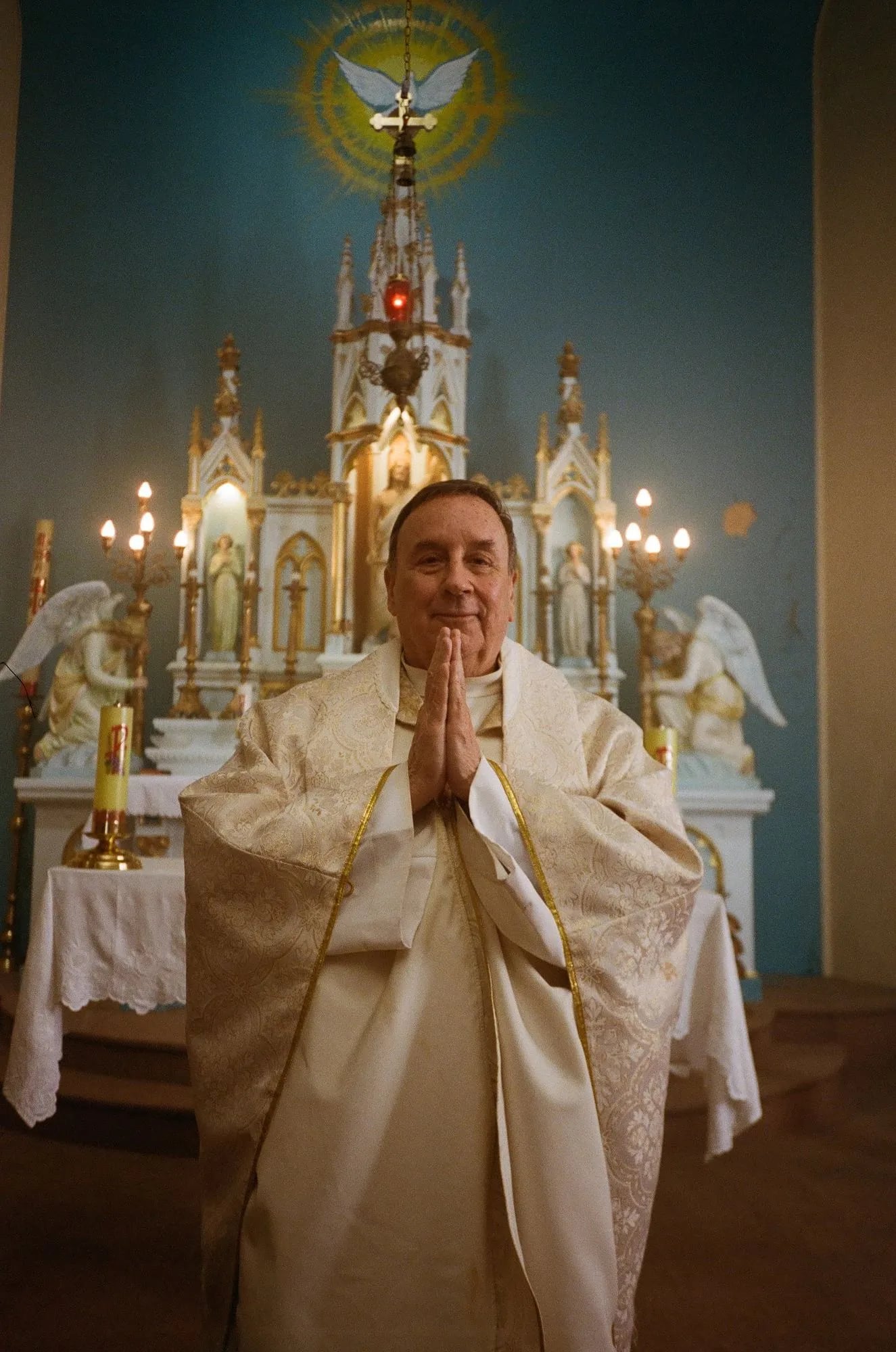 Smiling priest in cream vestments, hands pressed together, before an ornate white altar with candles and angel statues