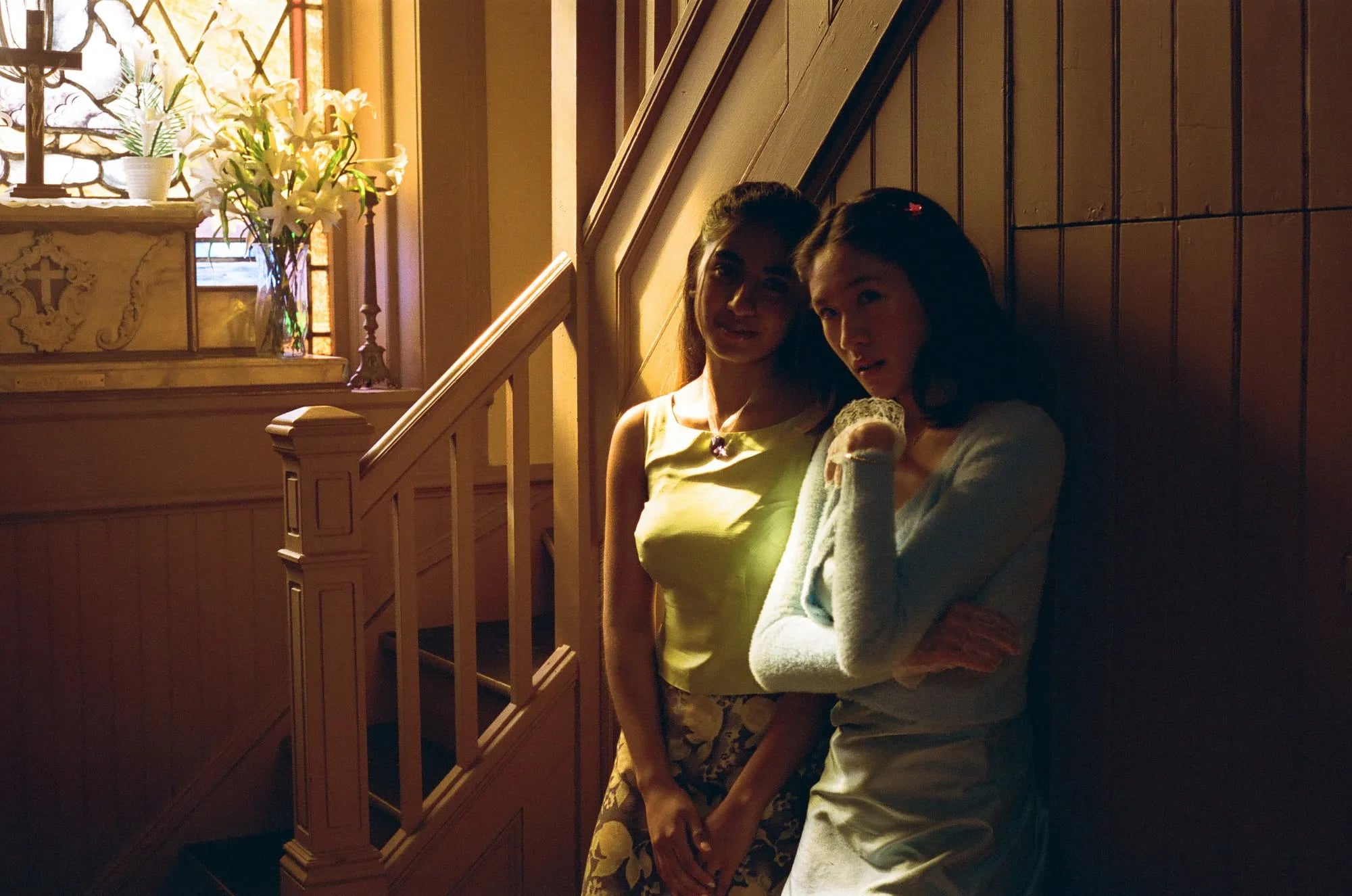 Two young women in pastel tops standing against wood paneling beside a staircase, with a cross and lilies in a window