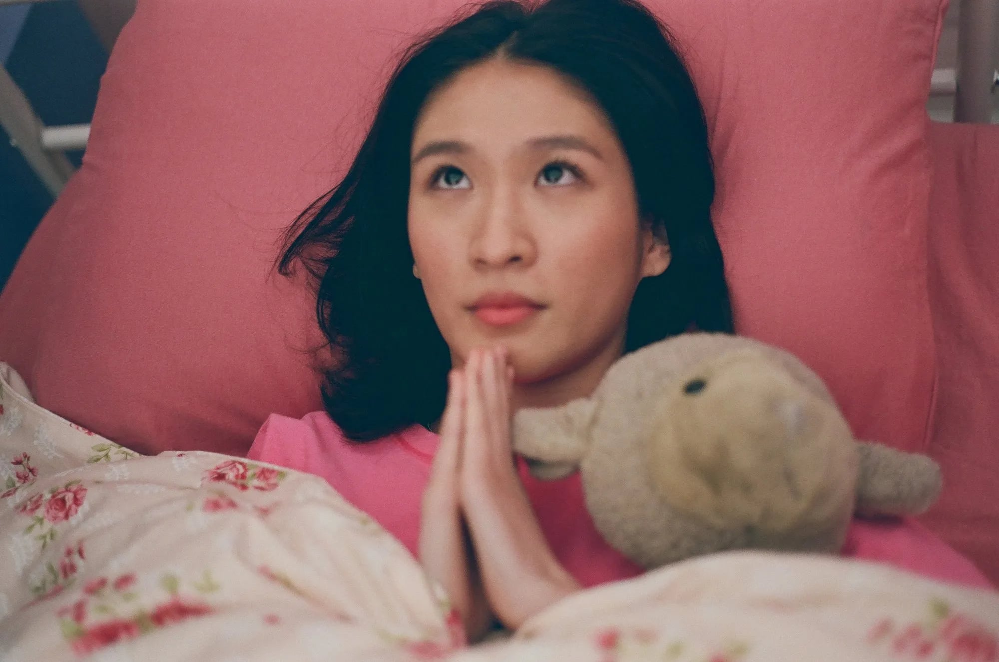 Frame from No Other Gods But Me: a young woman lying on a pink pillow, hands pressed in prayer, a stuffed lamb beside her