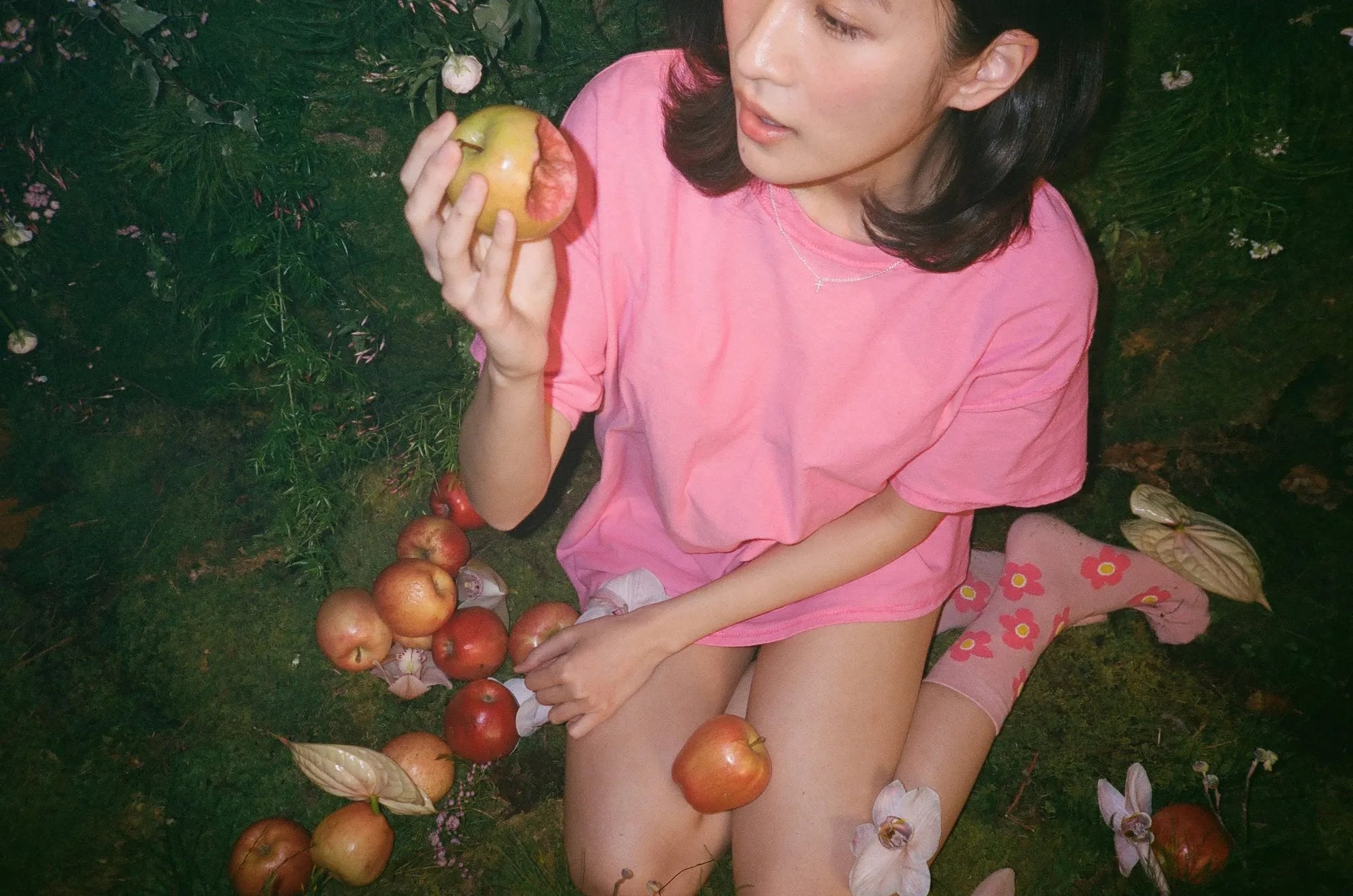Woman in a pink tee kneeling on moss, holding a bitten apple, with more apples and flowers scattered around her