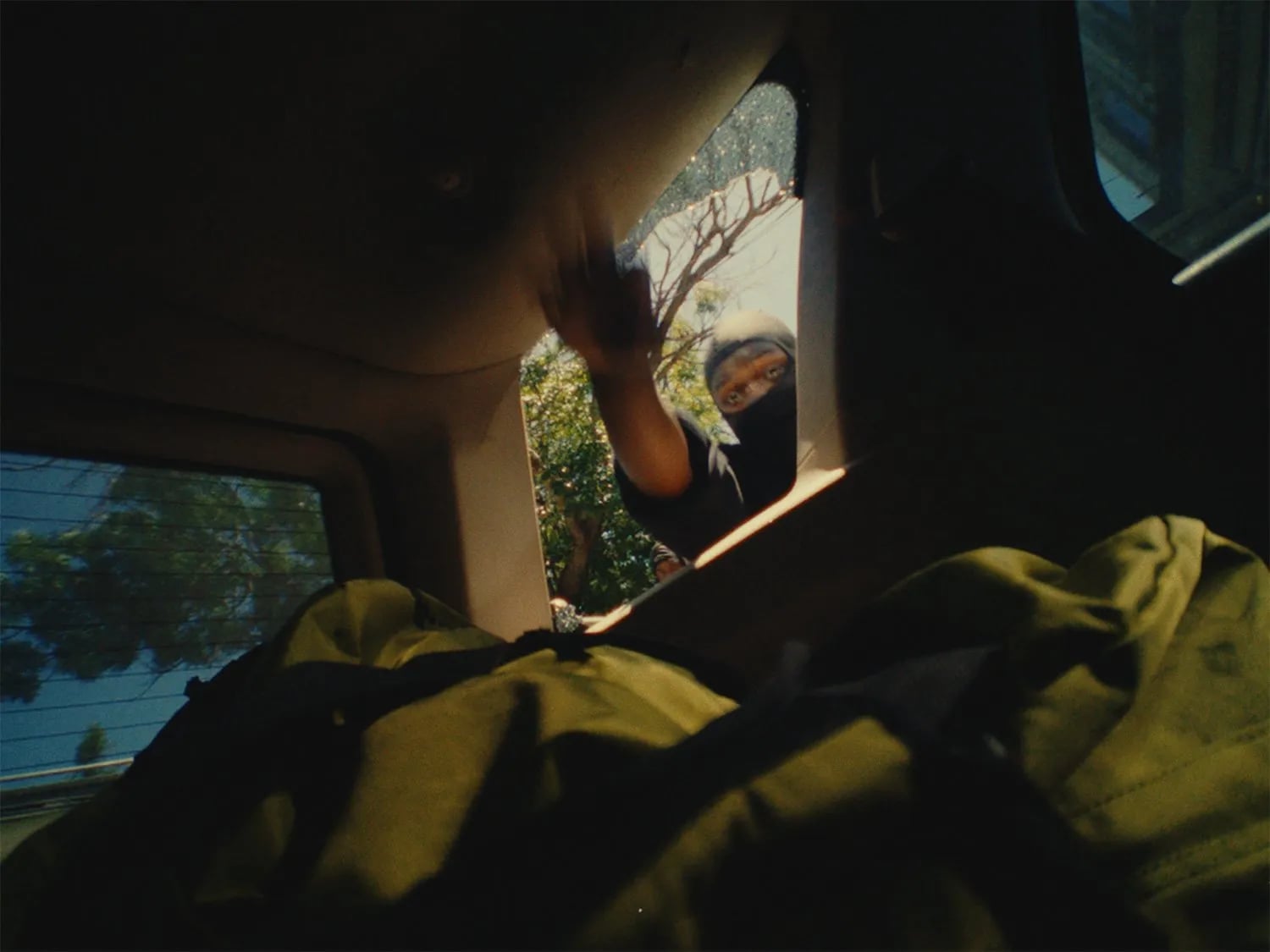 View from inside a car's cargo area toward a person in a black balaclava reaching for a bag through a broken rear window