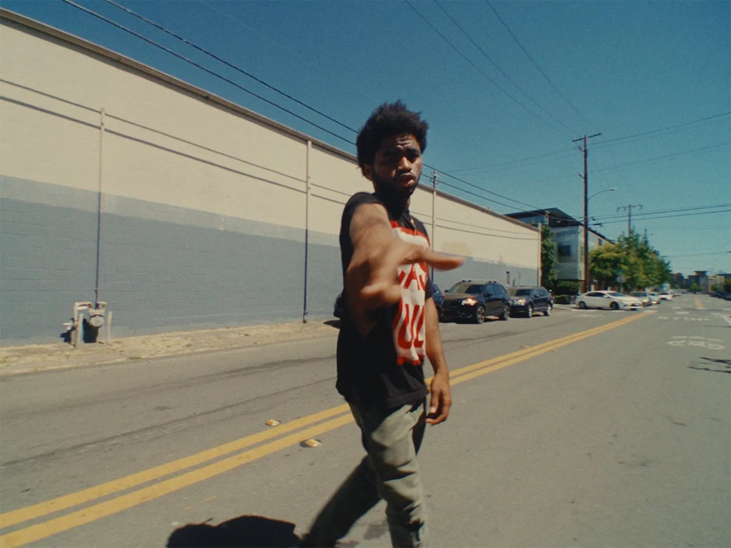 Man in a black tee reaching a hand toward the camera on a street with yellow lines, a long beige wall and blue sky behind