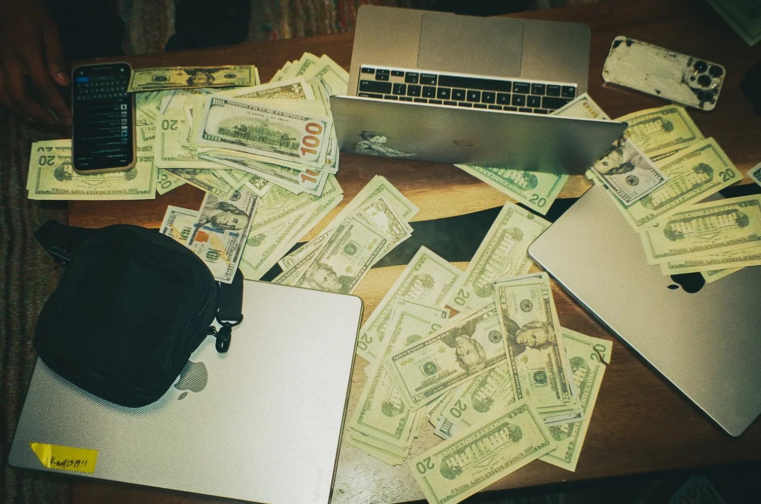 Bundles of twenty and hundred dollar bills spread across a wooden table with three laptops, two phones and a black bag