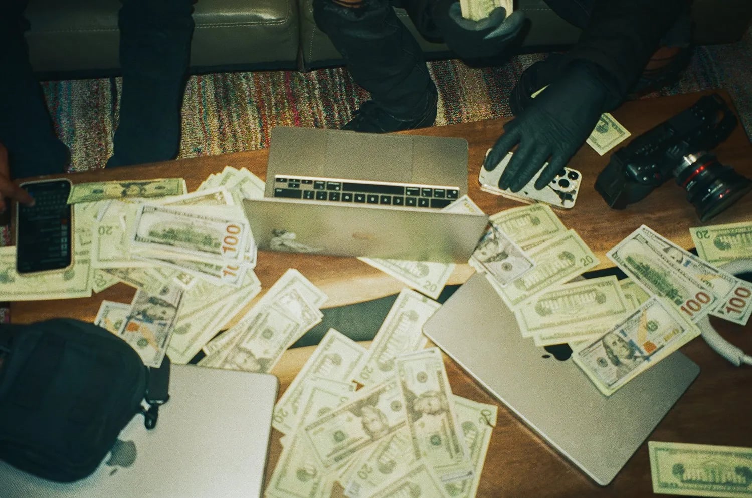 Overhead close-up of cash, laptops and phones on a wooden table, with gloved hands and a camera at the edge of the frame