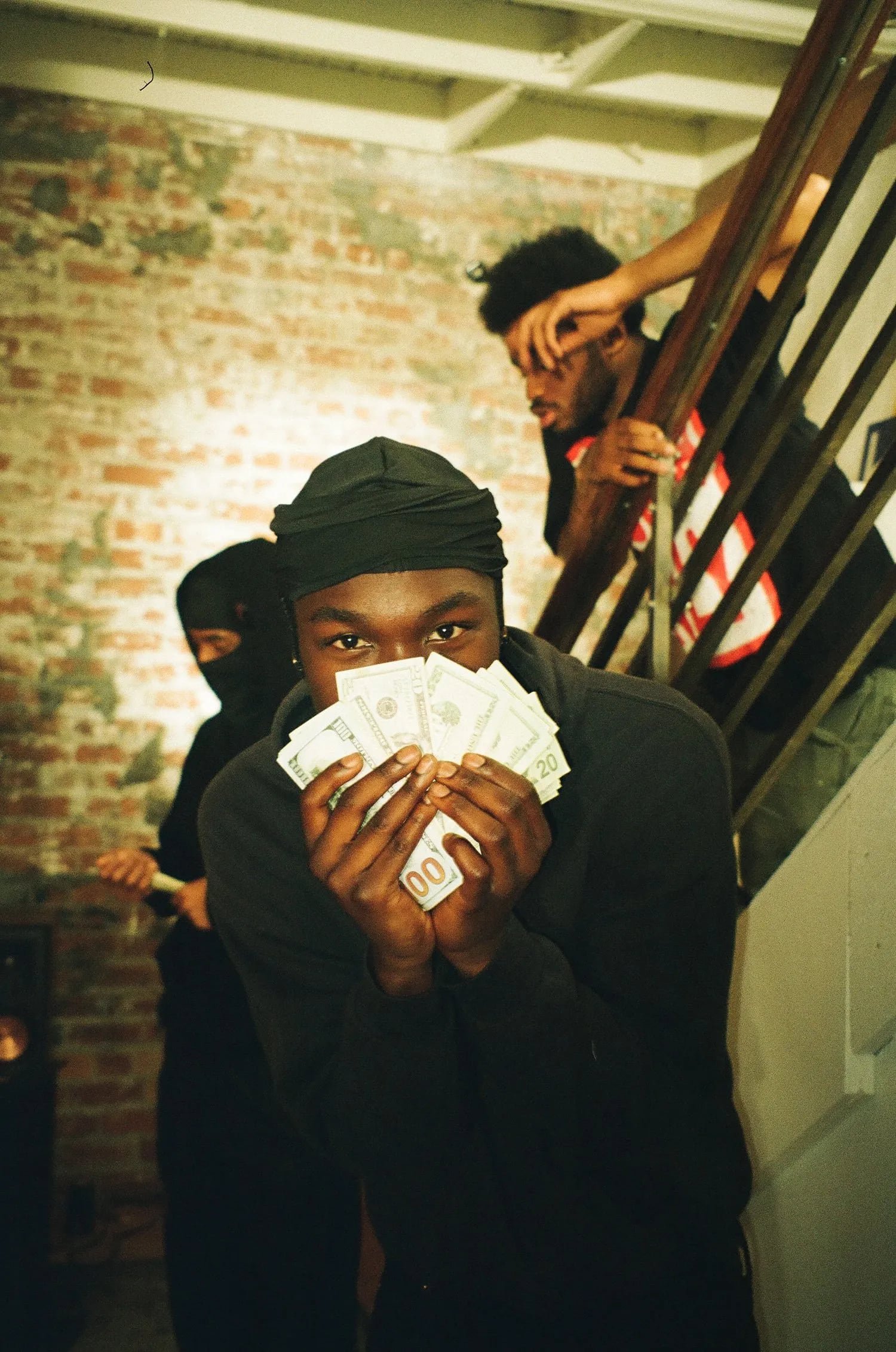 Man holding a fan of cash over his lower face in front of a brick wall, with a masked person and a man on a stairway behind