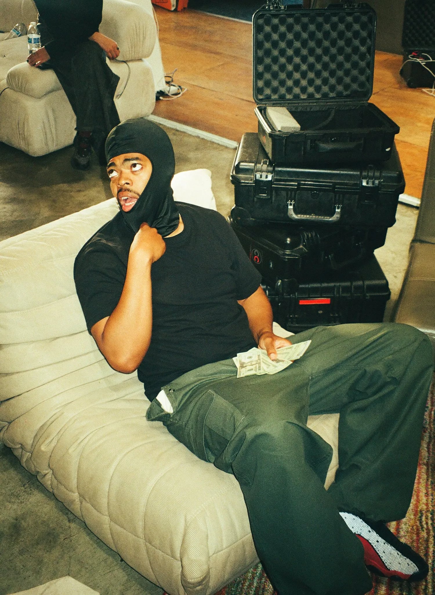 Man in a black tee with a balaclava pulled down on his face, sitting on a cream couch holding cash beside stacked black cases