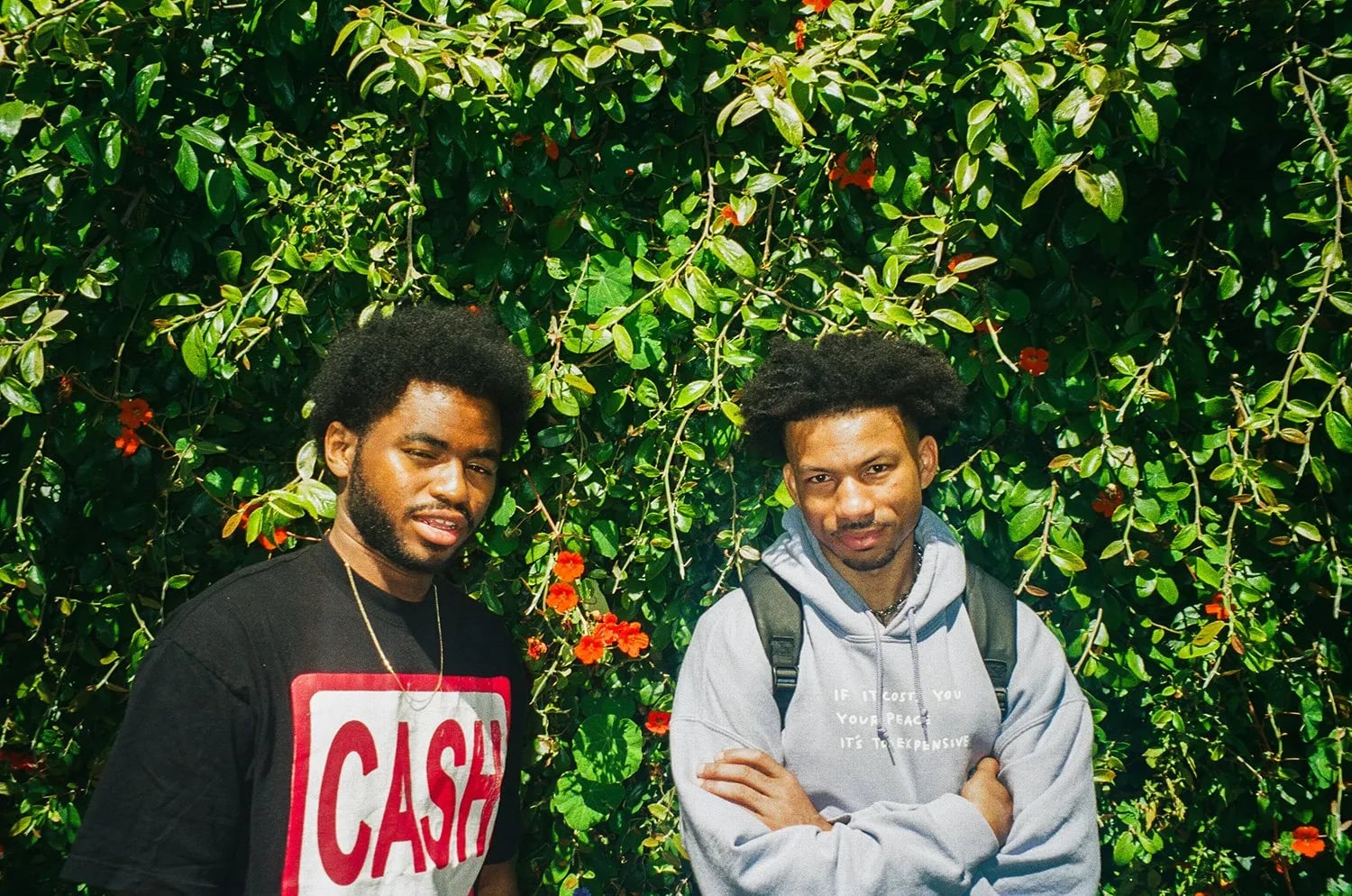 Two men in front of a green hedge with orange flowers, one in a CASH ONLY tee, the other in a grey hoodie and backpack
