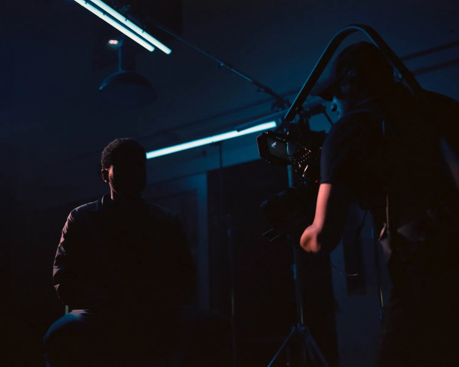 Camera operator with a rig faces a silhouetted man seated in a dark room lit by blue tube lights