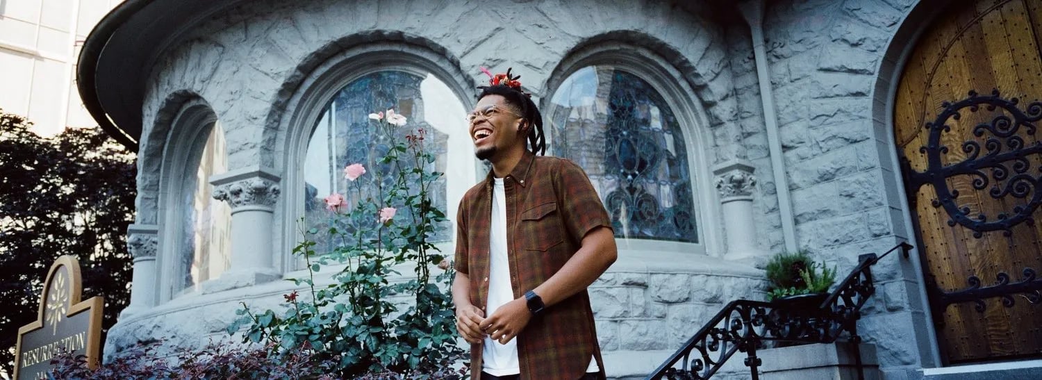 Ovrkast. Face video still: laughing man with dreads in a plaid shirt before a stone church