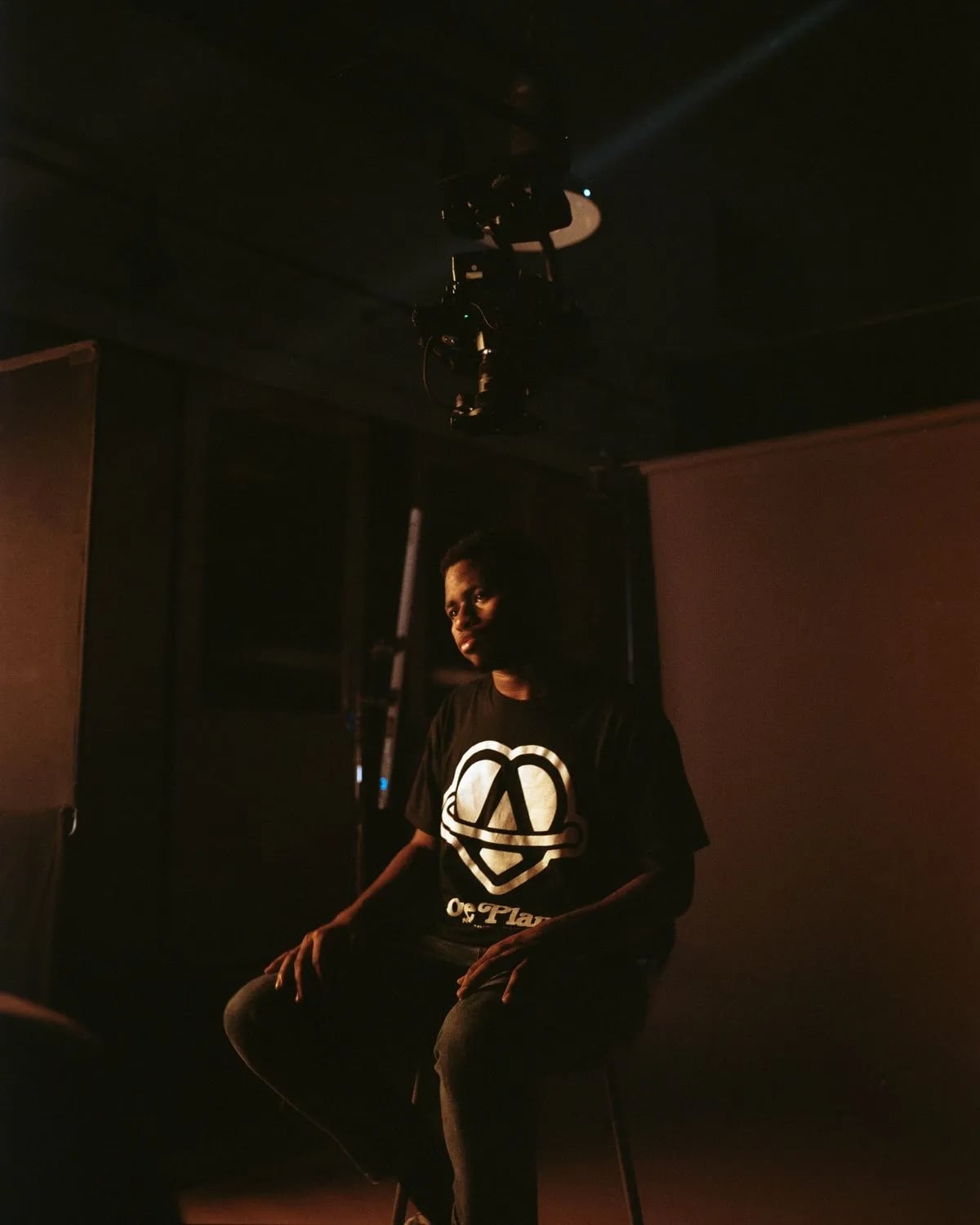 Man in a black graphic tee sits on a stool in a dark, warm-lit studio with a camera rig hanging overhead