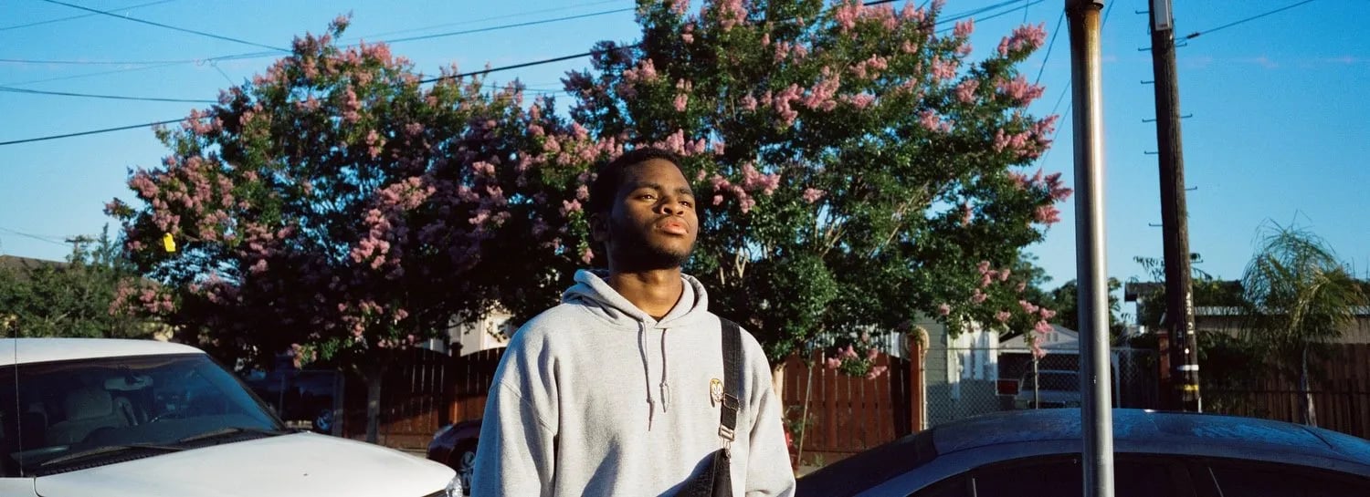 Man in a grey hoodie with a shoulder bag stands between parked cars in front of pink flowering trees