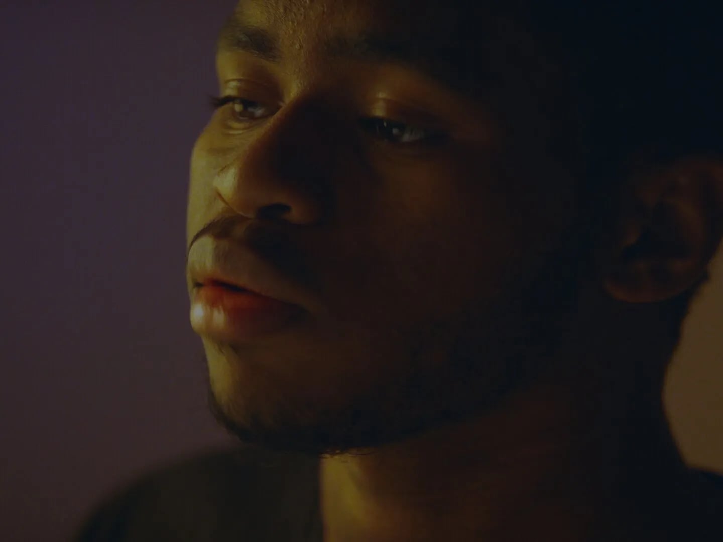 Frame from the Ovrkast. Face music video: close-up of a man's face half lit in warm, purple orange dim light, eyes lowered