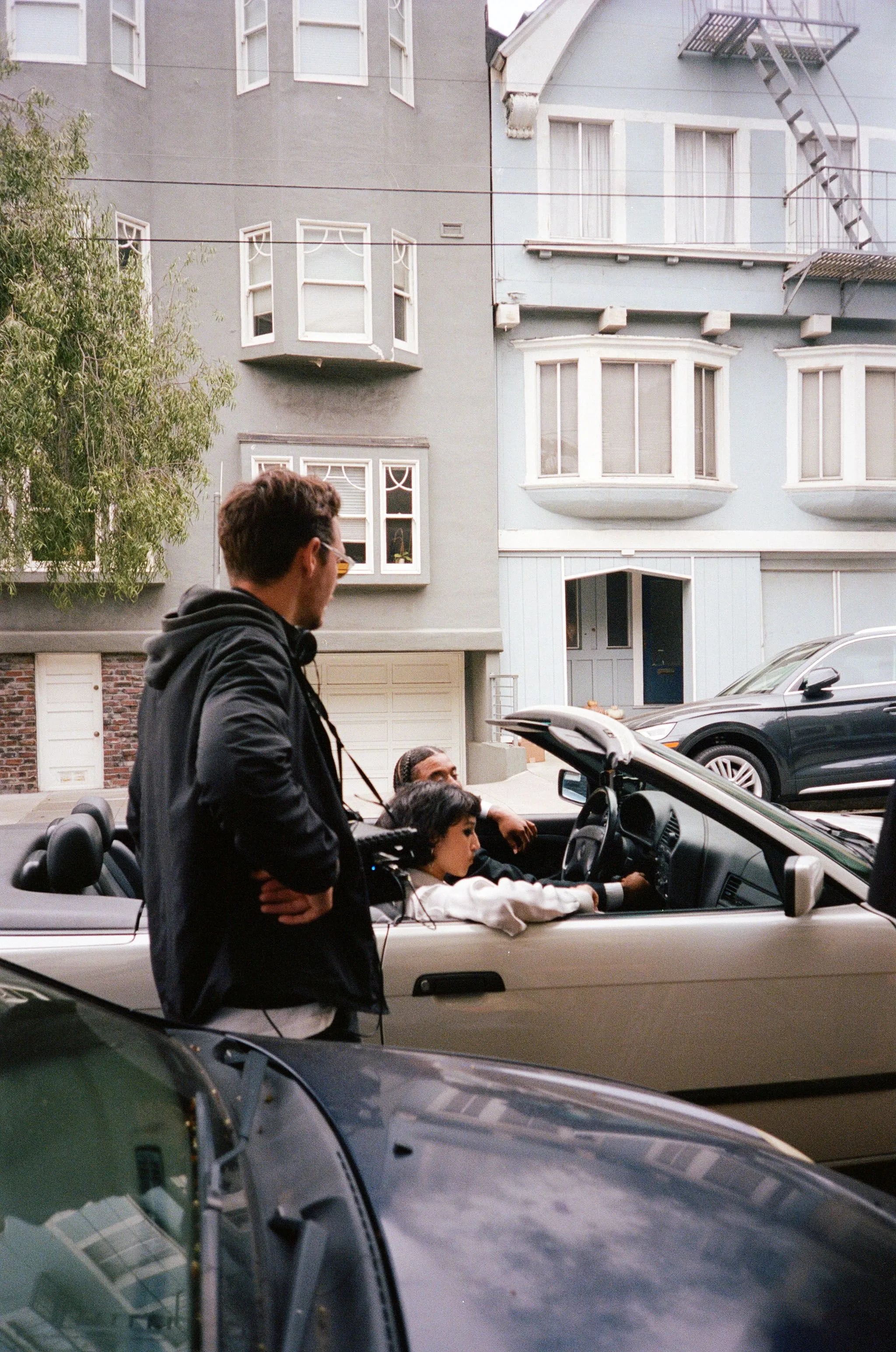 Skyler in a black hooded jacket wearing a director's monitor stands next to a light-colored convertible with two people in it
