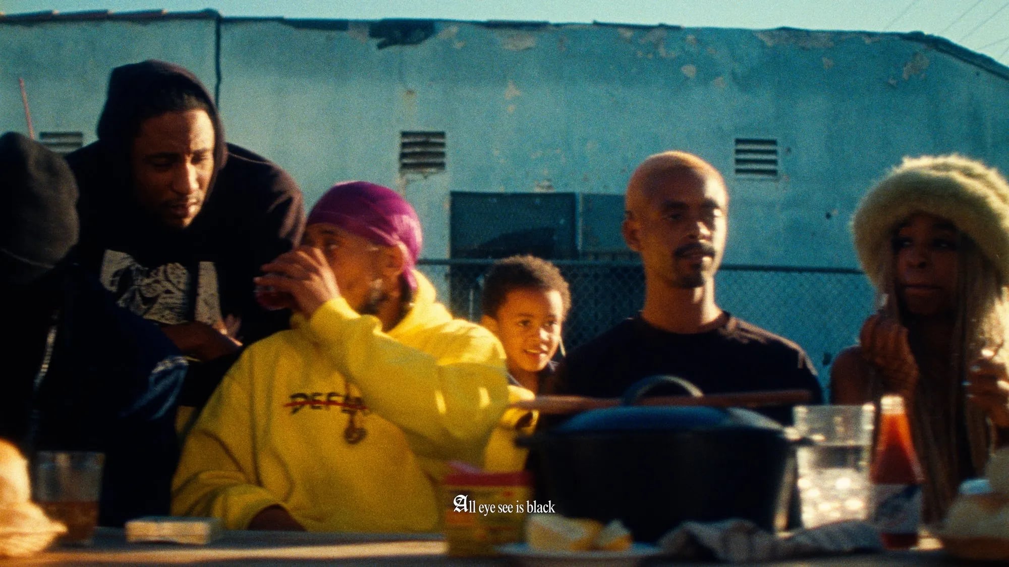 Group seated at an outdoor table in sunlight, a child behind them, subtitle "All eye see is black"