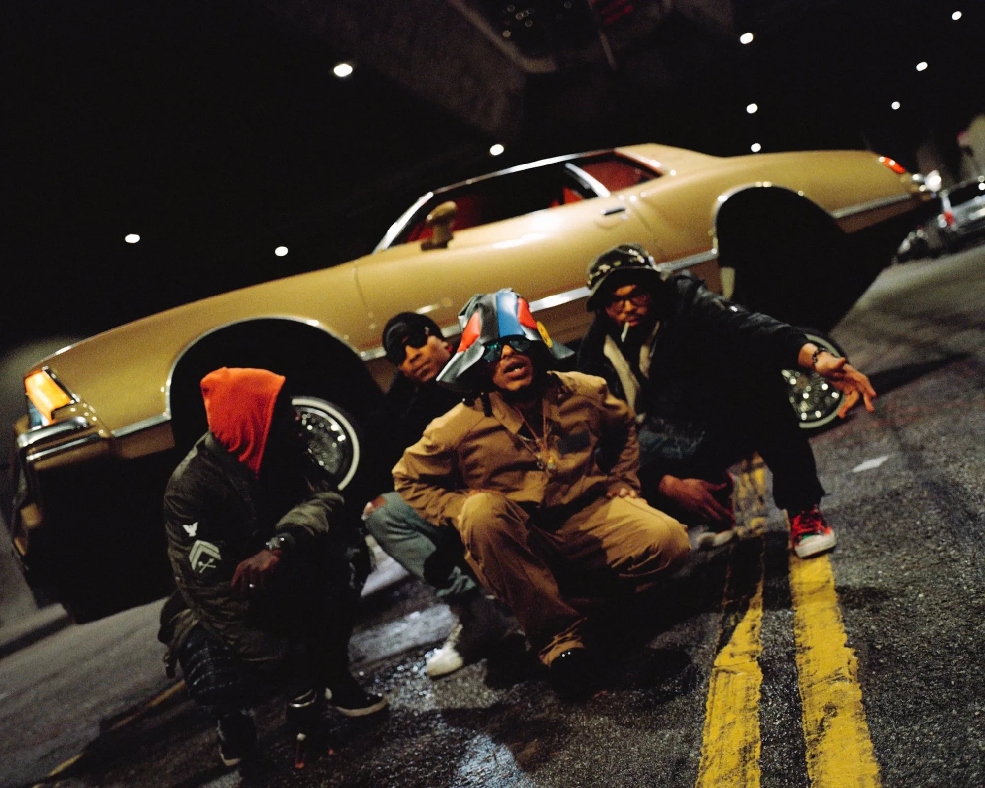 Four men crouching in a night tunnel in front of a tan lowrider, one in a colorful bucket hat, another smoking