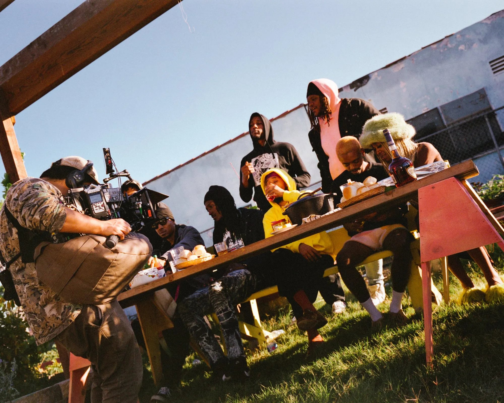 Behind the scenes: camera operator with a shoulder rig filming a group seated at a long outdoor table in a garden