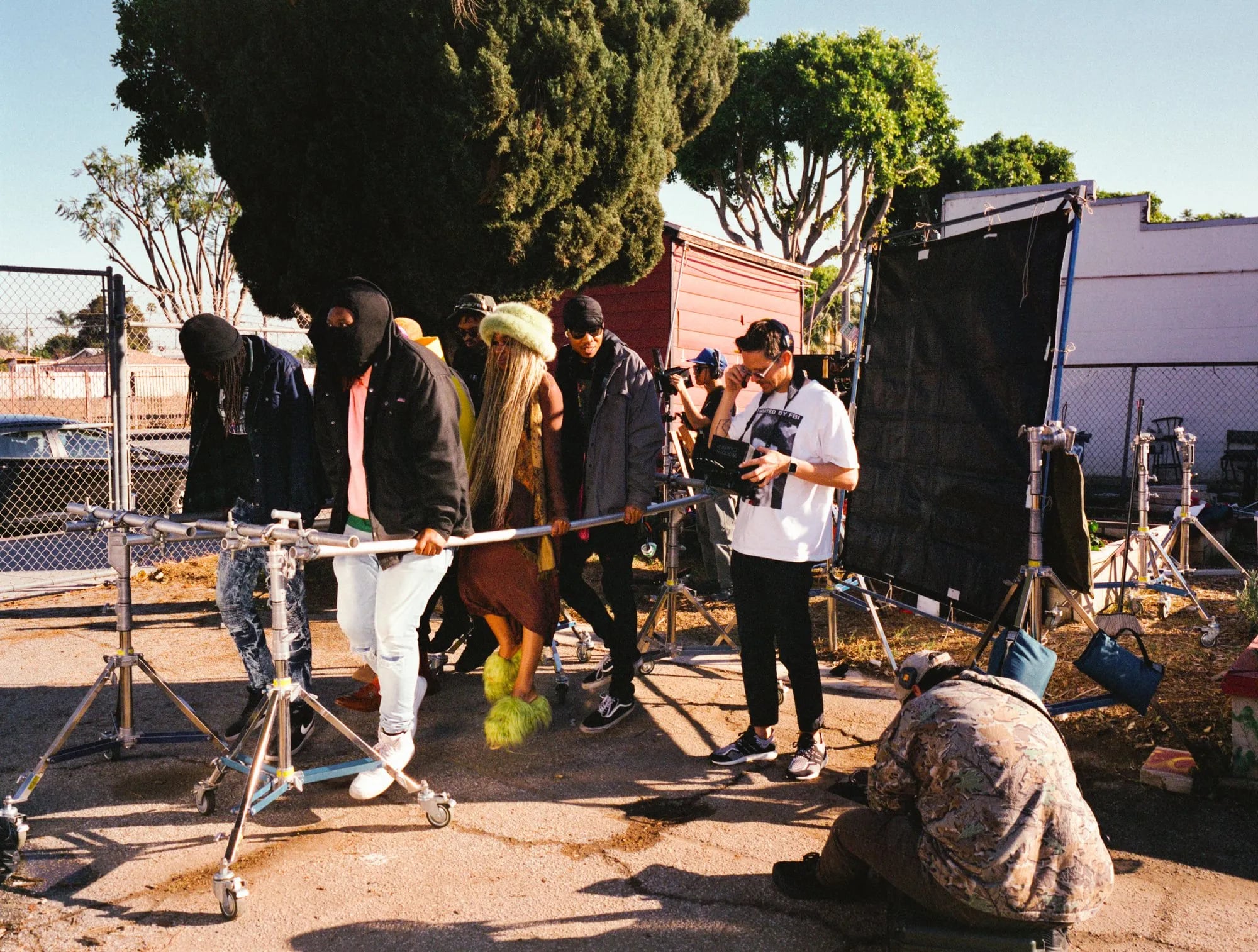 BTS: Cast holding themselves off ground on a raised pole across a lot, with a black flag and lighting stands behind