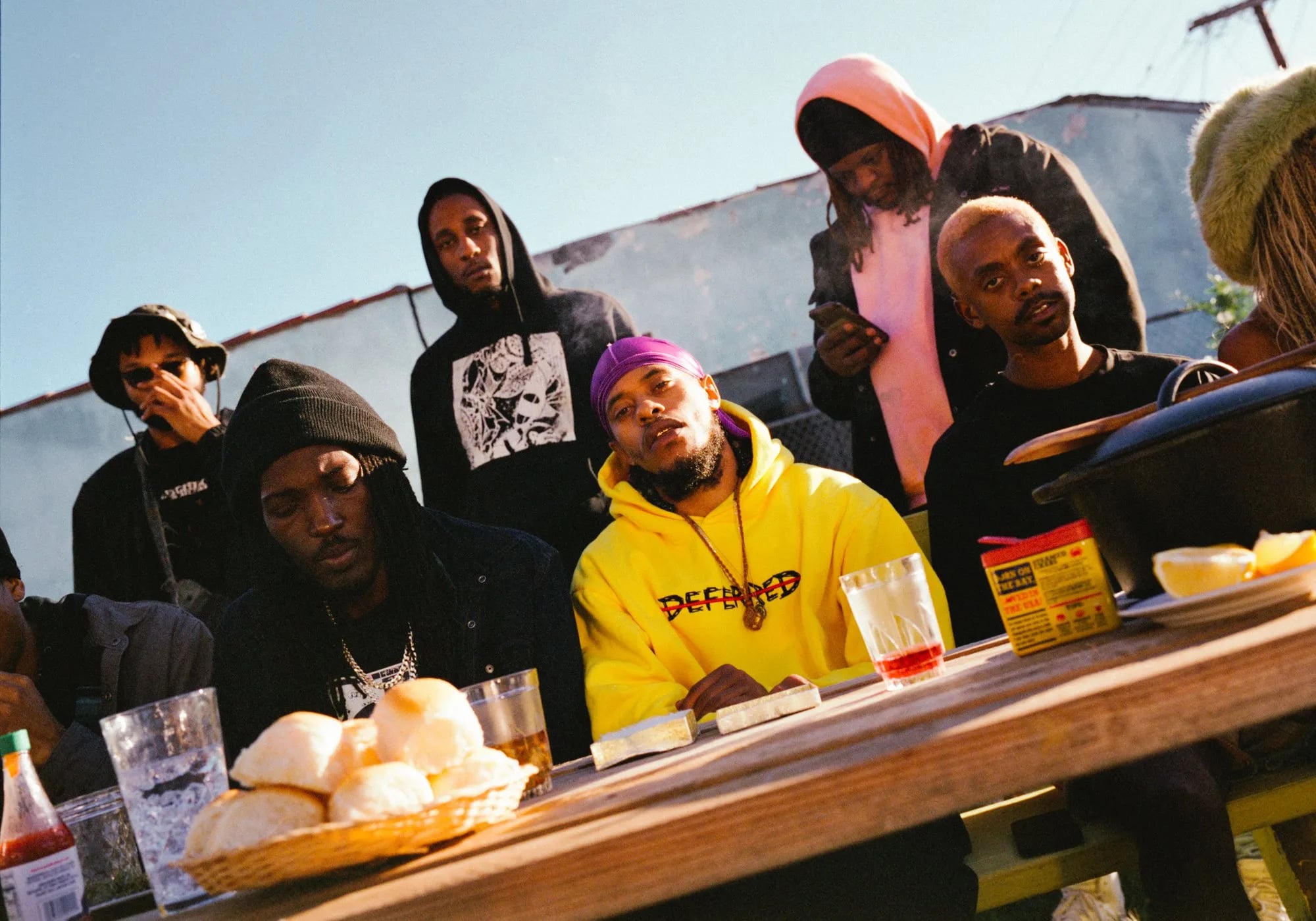 Group at an outdoor table, a man in a yellow hoodie and purple durag at centre, bread rolls and glasses in front