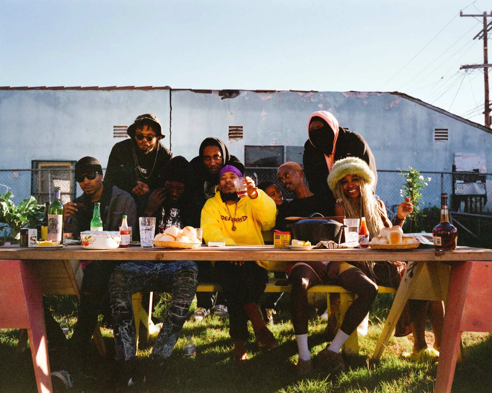 Group portrait around a long picnic table in a garden, a man in a yellow hoodie raising a cup, bottles and bread on it