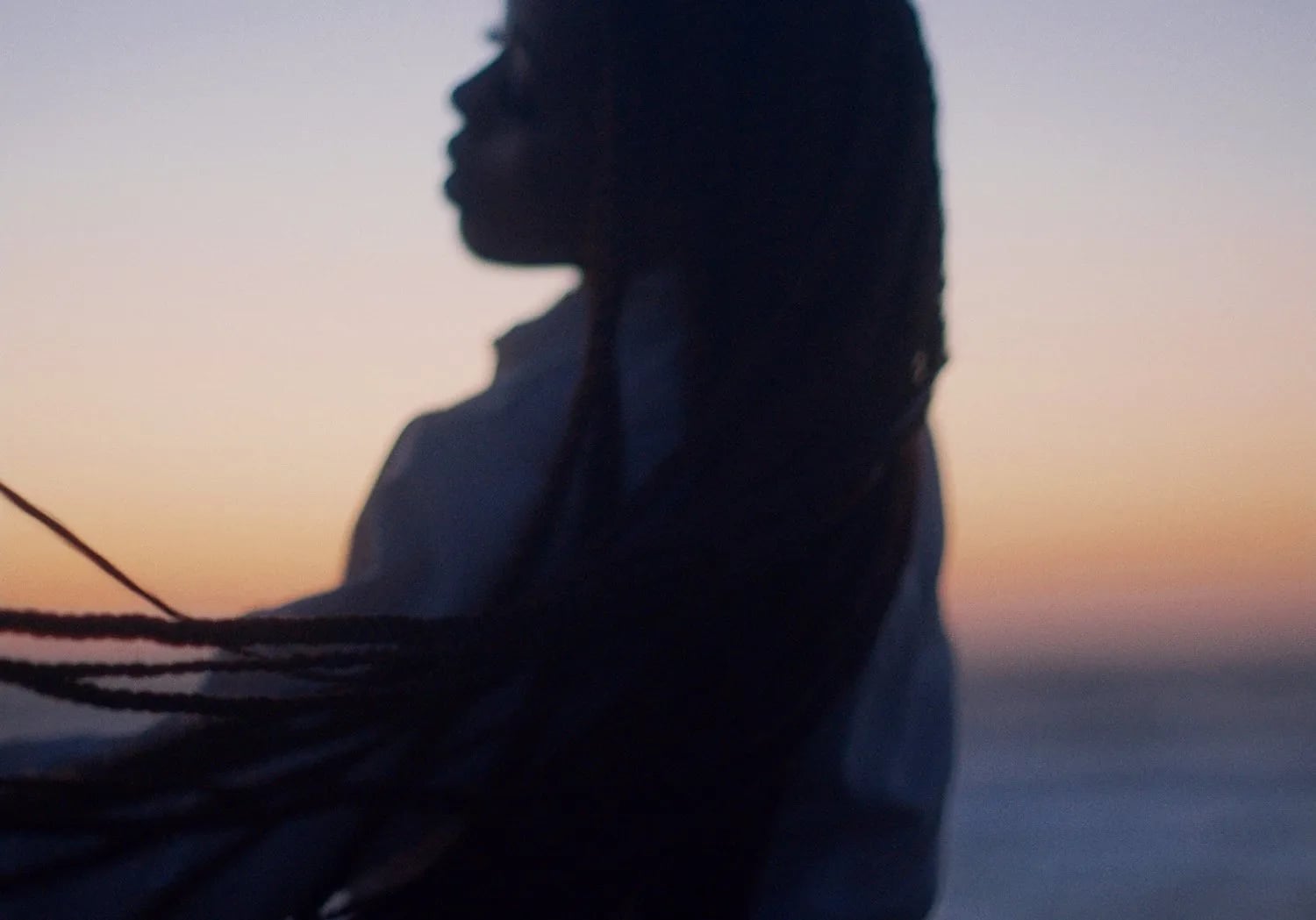 Blurred silhouette of a woman with long braids against an orange and violet sunset sky above the sea