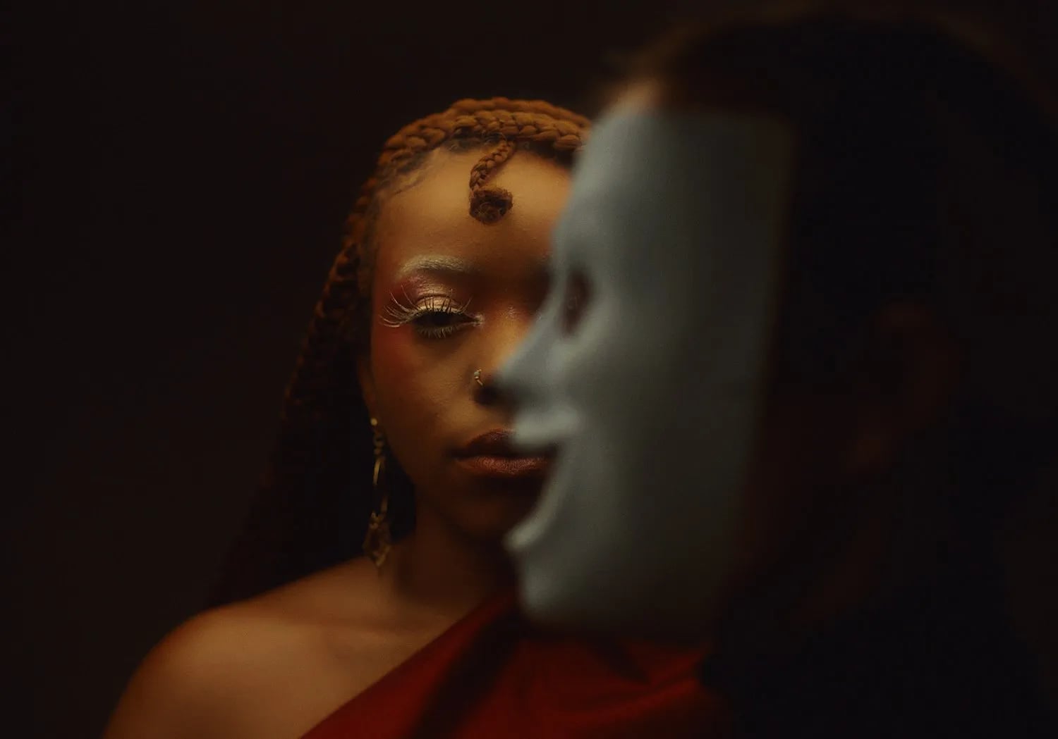 Close-up of a woman with braids and glittery lashes looking past a blurred white mask in the foreground