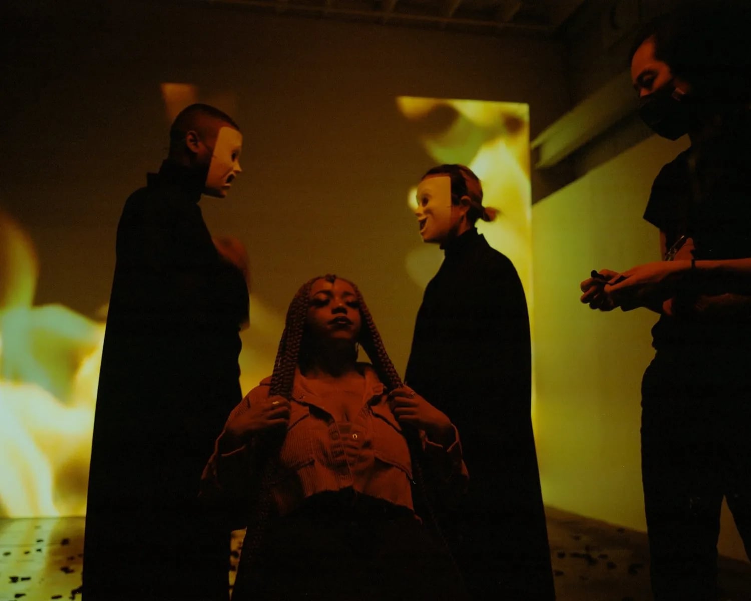 Woman with braids between two people in white masks in a dim room lit by projected flames, a masked figure on her left and right