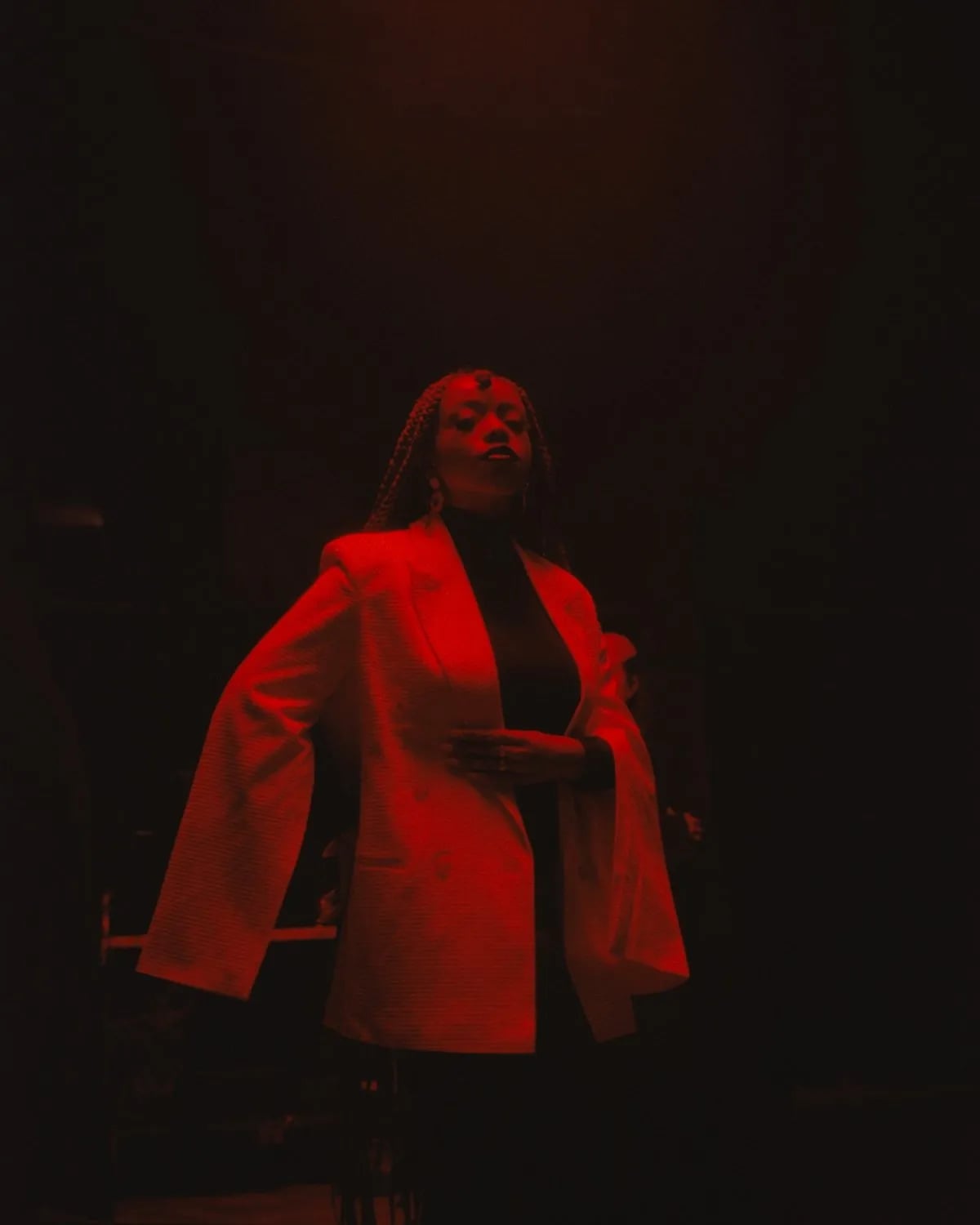 Woman with braids in a white cape-sleeved blazer standing in a dark room under deep red light, one hand on her chest