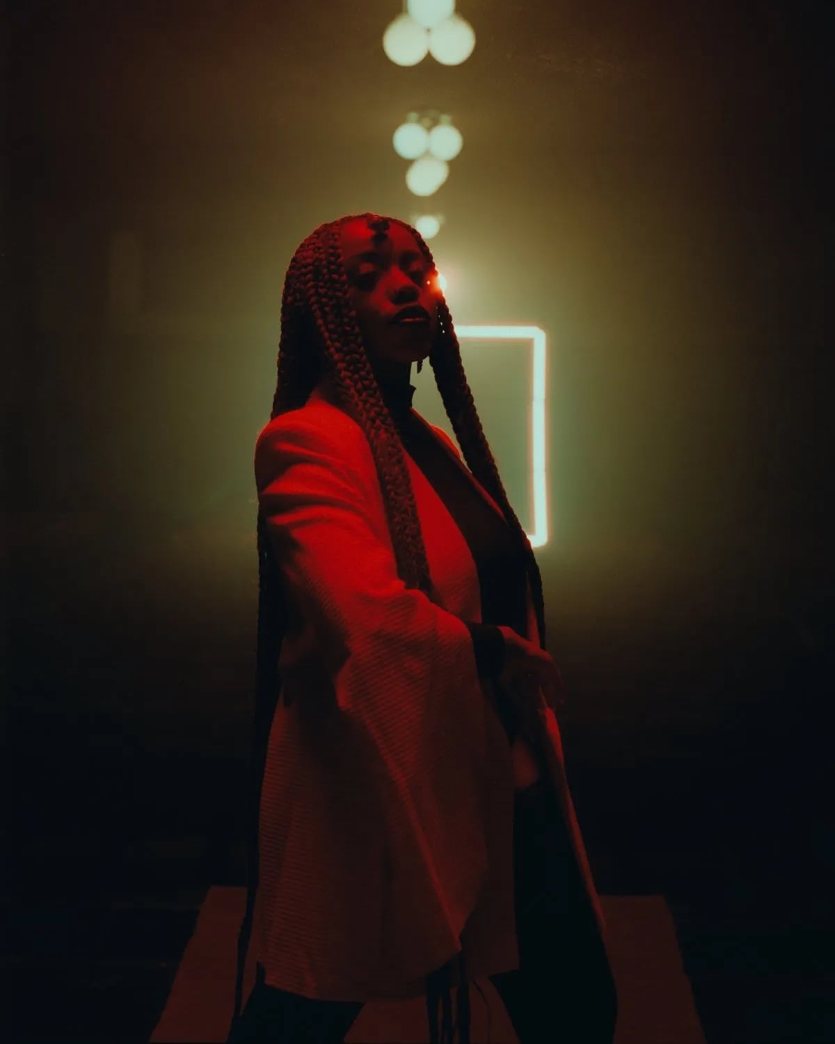 Woman with long braids in a white jacket lit red, standing in a dark hazy room with a glowing rectangle behind her