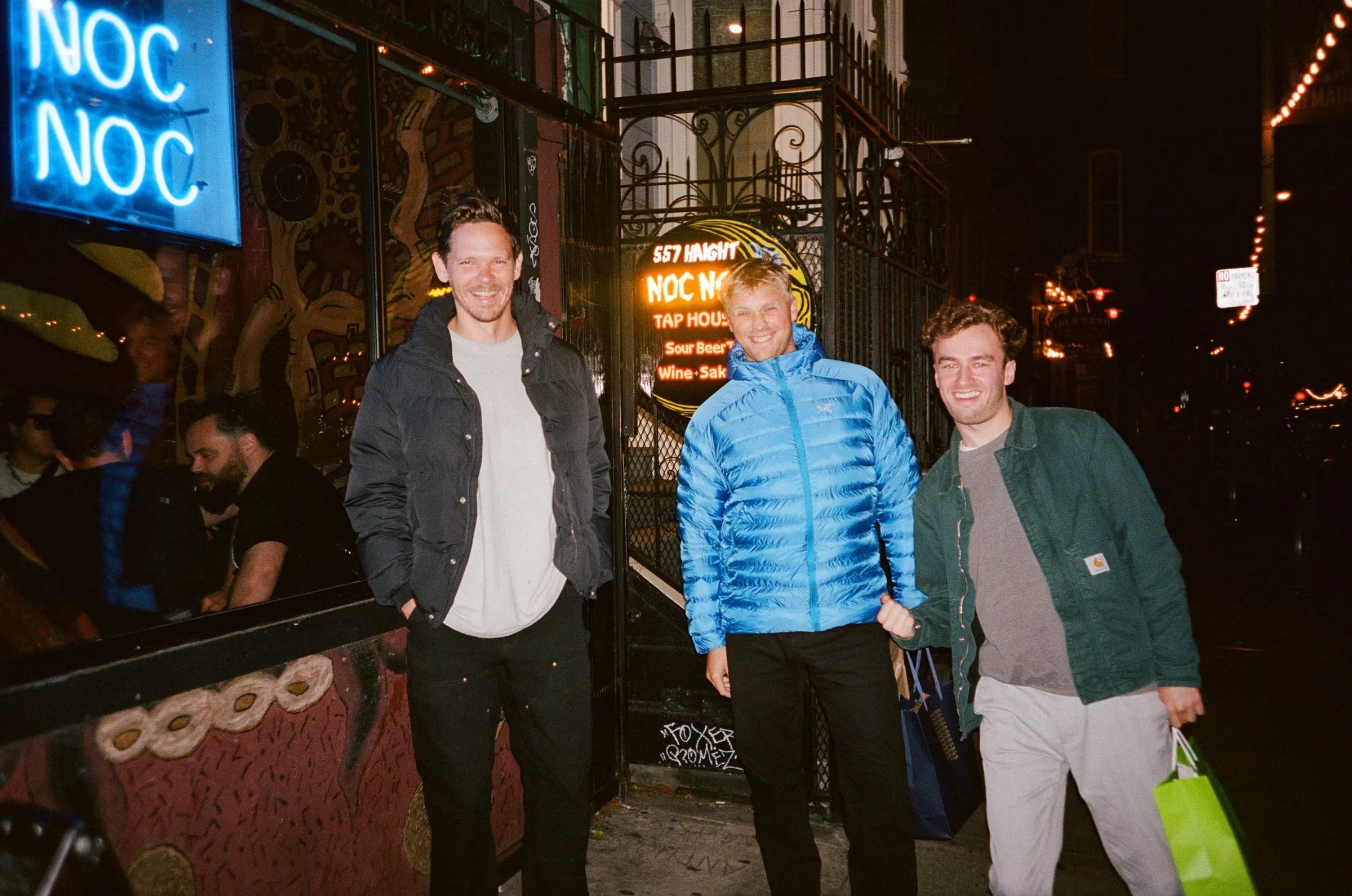 Three people smiling outside a bar at night under a blue neon NOC NOC sign, one in a bright blue puffer jacket