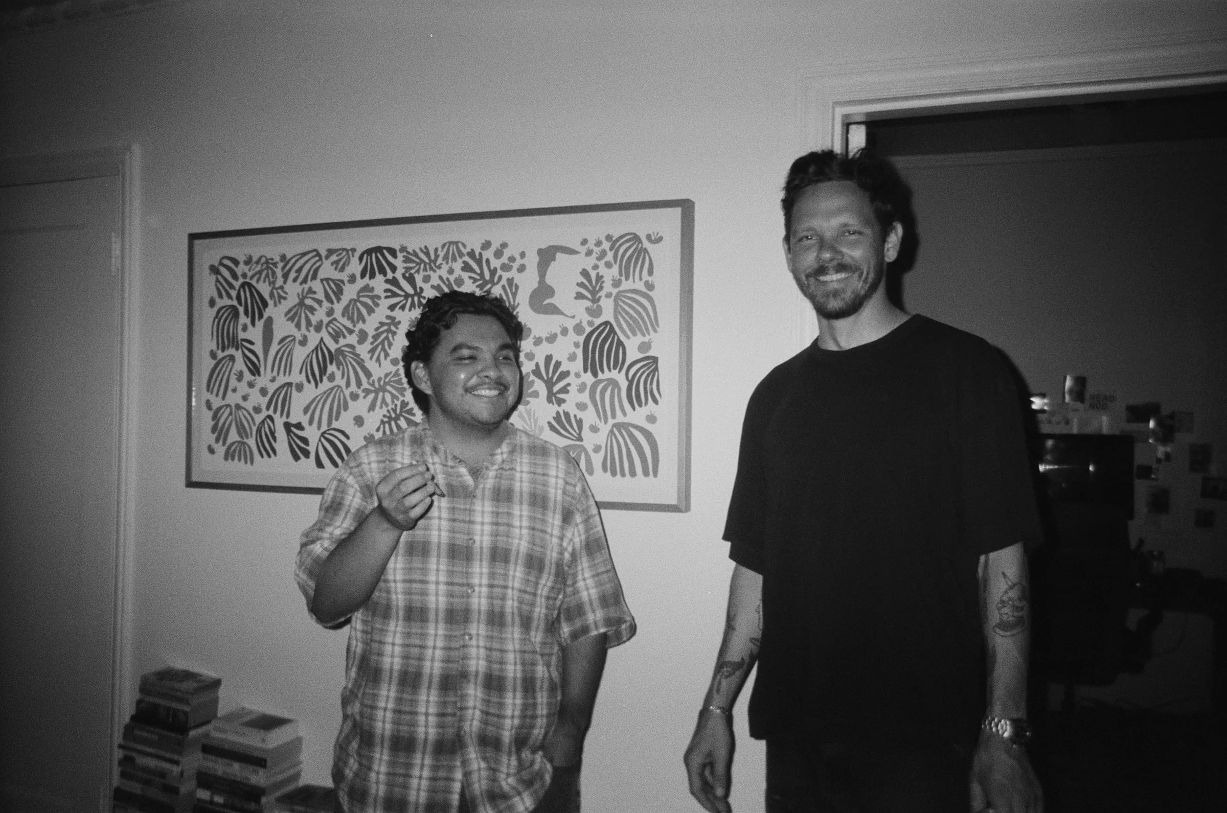 Skyler and a man smiling standing in front of a framed leaf-pattern print on a wall, black and white film
