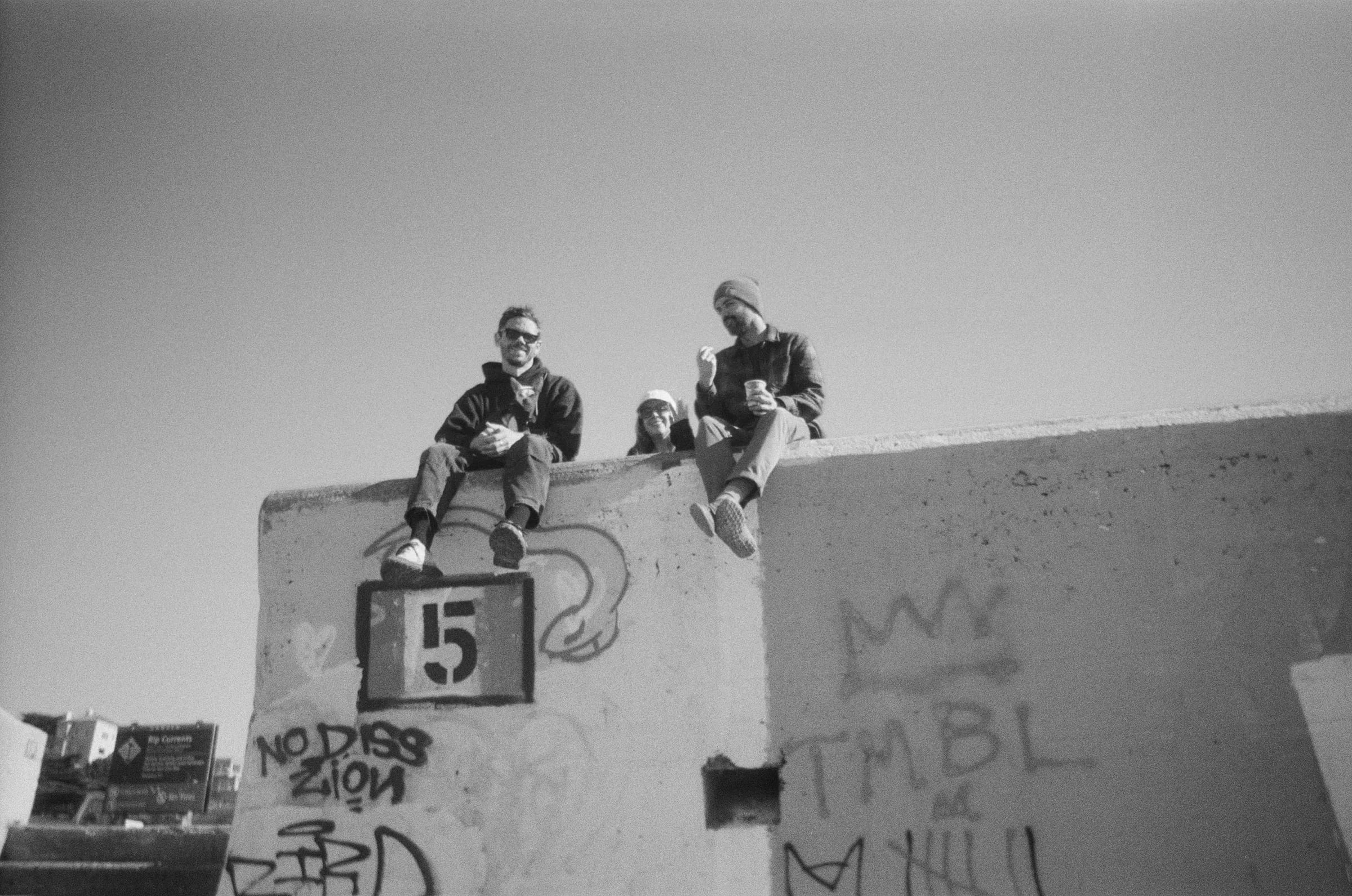 Skyler and two people sitting on top of a graffiti-covered concrete wall under a clear sky, black and white film