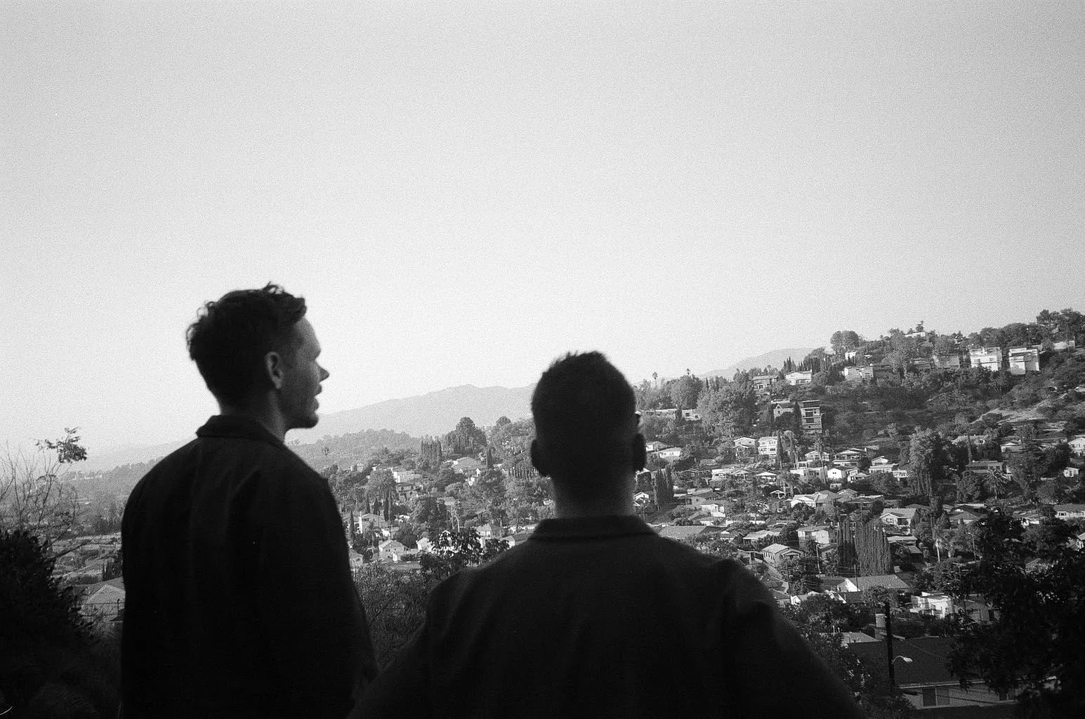 Two people seen from behind looking out over a hillside of houses and distant mountains, black and white